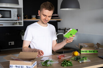 Happy Caucasian white man Unpack Online Home Food Delivery. Box with packed tuna, shrimp, vegetables and recipe card on a kitchen background. Food delivery services. Dinner Set from restaurant