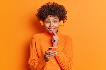 Pretty satisfied African American woman smiles gently at camera keeps finger near lips uses...