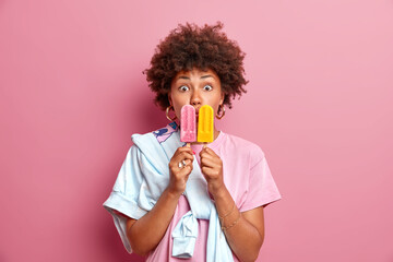 Stunned curly haired ethnic teenage girl covers mouth with two frozen ice creams has sweet tooth enjoys eating yummy summer dessert wears casual t shit and jumper isolated over pink background