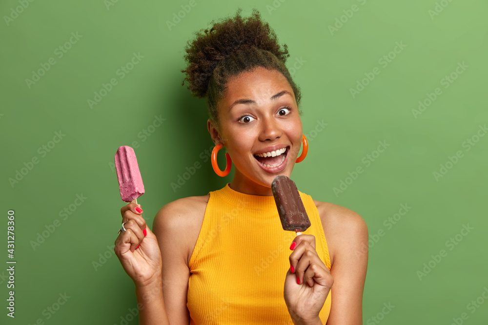 Wall mural joyful dark skinned young woman with combed curly hair has fun eats tasty ice creams of chocolate an