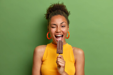 People unhealthy junk food and summer time concept. Cheerful dark skinned woman winks eye eats delicious chocolate ice cream has fun wears casual bright clothes isolated over green background