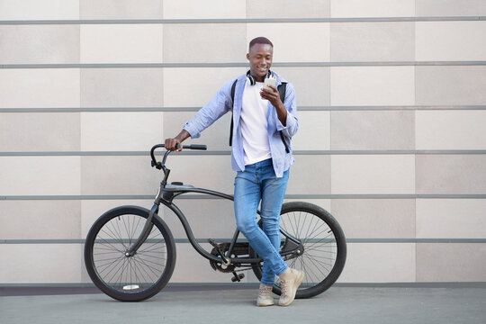 Cheerful African American man with bicycle standing near brick wall and choosing music playlist on smartphone