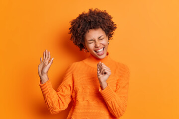 Positive dark skinned young woman with curly hair sings song loudly keeps hand near mouth as if microphone foolishes around wear casual jumper isolated over orange background. Monochrome shot