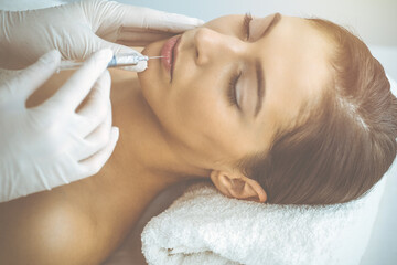 Obraz na płótnie Canvas Beautician doing beauty procedure with syringe to face of young brunette woman in sunny clinic. Cosmetic medicine and surgery, beauty injections