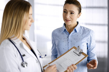 Young woman-doctor and her patient are discussing patient's current health examination, while...