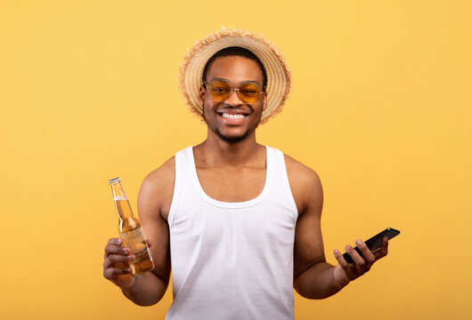 Joyful African American guy holding bottle of beer and smartphone on yellow studio background