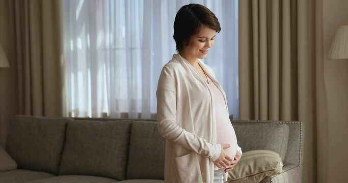 Young happy pregnant woman standing alone in cozy living room at home hold hands on belly stroking unborn child in tummy express love and protection smile look at camera. Maternity, pregnancy concept