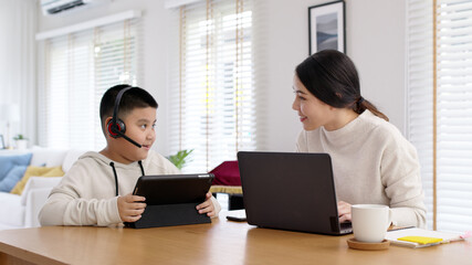 Young adult asian mother and kid stay home work and learning online remotely from computer tablet in self isolate from lockdown coronavirus covid19 social distance. Homeschool boy with parent.