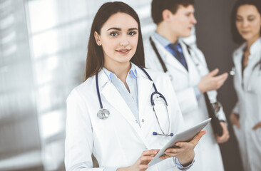 Smiling intelligent woman-doctor is holding a tablet computer in her hands, while she is standing together with her colleagues in a clinic. Physicians at work. Perfect medical service in a hospital