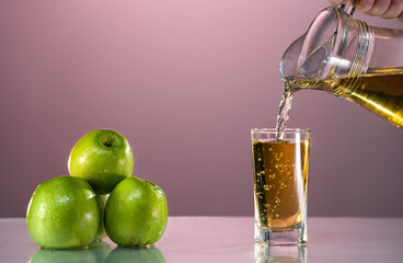 Fresh apple juice is poured from a carafe into a glass. Nearby are green apples on the reflective...