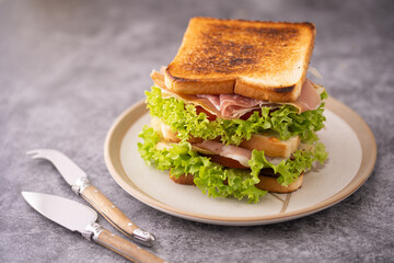 Big toast sandwich with lettuce, ham, tomatoes and cheese and two cheese knives on grey surface