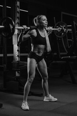 Fototapeta na wymiar A sporty woman with blonde hair is started squatting with a barbell near the squat rack in a gym. A girl is doing a leg workout.