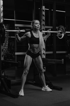 A sporty woman with blonde hair is squatting with a barbell near the squat rack in a gym. A girl is doing a leg workout.