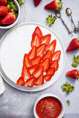White cheesecake with sliced strawberries surrounded by whole strawberries and strawberry jam on grey surface, flat-lay top view