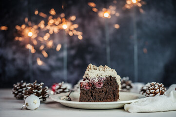 Fototapeta na wymiar A slice of chocolate cake with cherries and beige icing on a beige plate surrounded by christmas decoration and sparklers on the background