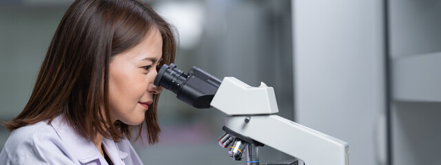 A young scientist woman in a laboratory coat looking through a microscope in a laboratory to do...