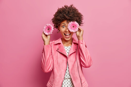Positive female florist covers eyes with fragrant gerbera flowers makes bouquets for special occasions smiles broadly wears fashionable jacket isolated over pink background. Feminity concept.