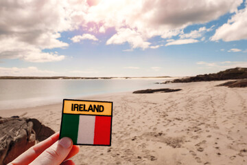 Badge with sign Ireland and National flag in focus. Gurteen beach out of focus, county Galway, Ireland. Warm sunny day. Cloudy sky. Sun flare. Tourists footprints on the sand