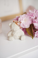A grey concrete figurine of a teddy bear on a white surface holding golden earrings and surrounded by pink cherry blossom