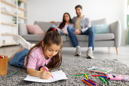 Family happiness. Cute girl drawing with colorful pencils, lying on floor carpet while parents relaxing on sofa