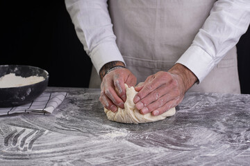 A male chef baker in a white shirt and grey apron kneading dough on a dark kitchen table