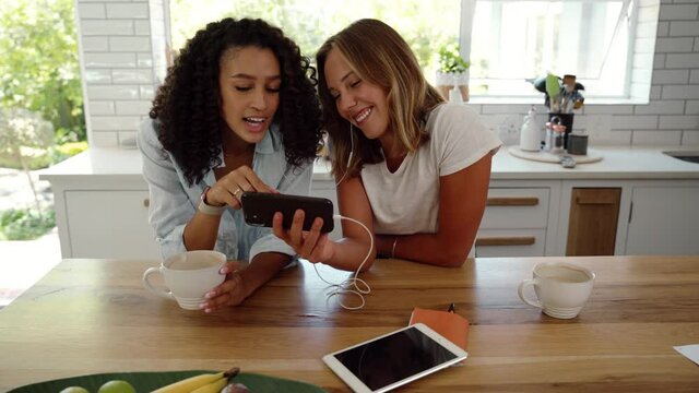 Mixed race female friends holding cellular device chatting in kitchen scrolling through images 