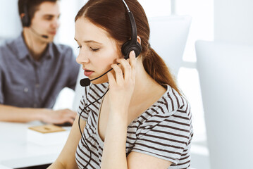 Casual dressed young woman using headset and computer while talking with customers online. Group of operators at work. Call center, business concept