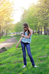 A sad young girl stands in a park against a backdrop of green trees and a sunset in sunglasses, a blue plaid blouse and jeans, and looks away. Full-length portrait. Vertical orientation.
