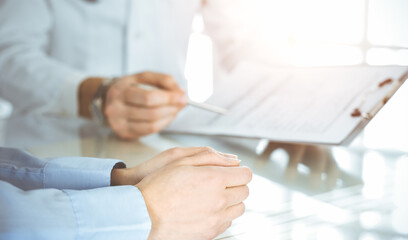 Obraz premium Unknown male doctor and woman-patient discussing current health examination while sitting in sunny clinic and using clipboard. Good medical service in hospital