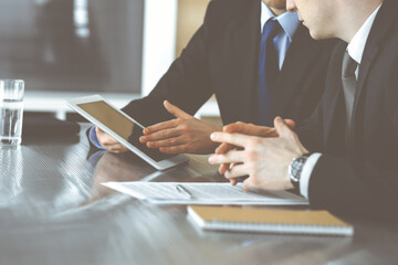 Unknown businessmen using tablet computer and work together at the glass desk in modern office, close-up. Teamwork and partnership concept