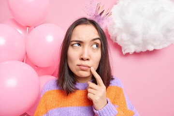 Lovely brunette woman being deep in thoughts keeps finger near lips daydreams about something wears striped sweater little crown holds bunch of inflated balloons isolated on pink studio wall