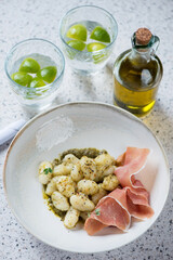 Beige plate with potato gnocchi, green basil pesto and prosciutto, vertical shot on a beige stone background, selective focus