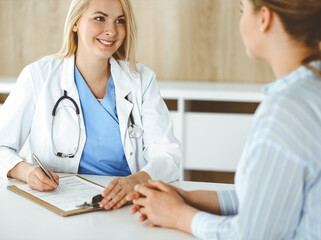 Woman-doctor and patient sitting and talking in hospital or clinic. Blonde therapist is cheerfully smiling. Medicine concept