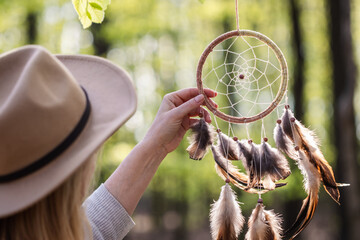 Woman with hat is hanging dreamcatcher in woodland. Indian ritual decoration. Atmospheric mood and spirituality concept - obrazy, fototapety, plakaty