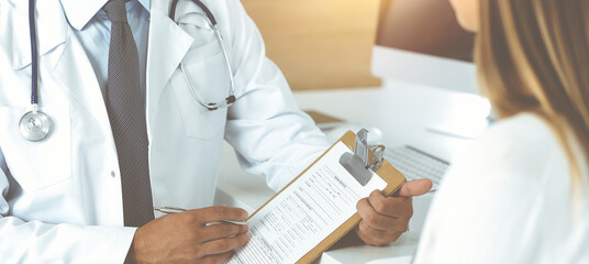 Doctor and patient discussing medical exam results while sitting at the desk in sunny clinic, close-up. Male physician using clipboard.