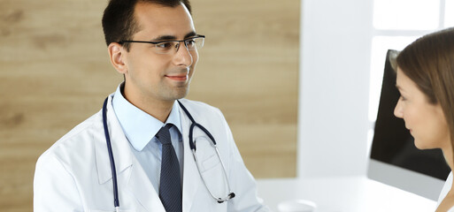 Doctor and patient discussing medical exam results while sitting at the desk in clinic. Male physician using tablet computer for filling up medication history record of young woman. Data in medicine