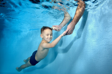A little boy swims under the water in the pool. Dad puts his hands under the water and catches the...