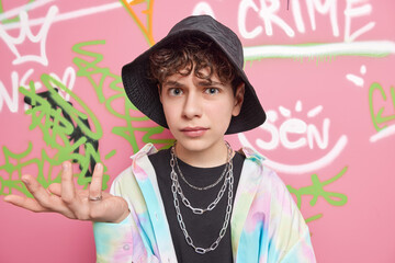 Cool looking indignant youngster shrugs shoulders and looks clueless at camera dressed in fashionable clothes poses against graffiti wall poses at urban place. Youth lifestyle and hooliganism