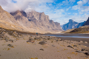 Hiker with a heavy backpack taking picture in remote arctic valley on a beautiful day of arctic summer. Dramatic arctic landscape of Akshayuk Pass, Baffin Island, Canada. Mt.Thor in the back