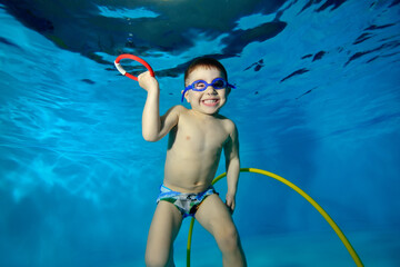 Funny little boy swims and poses underwater in the pool with a toy in his hand. He looks at the camera and smiles broadly. Portrait. Horizontal view. - obrazy, fototapety, plakaty