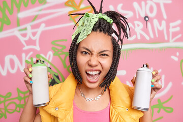 Modern teenage girl has dreadlocks looks with cheeky face expression at camera holds two aerosol bottles for drawing graffiti dressed in fashionable clothes. Millennial generation. Grunge style - obrazy, fototapety, plakaty