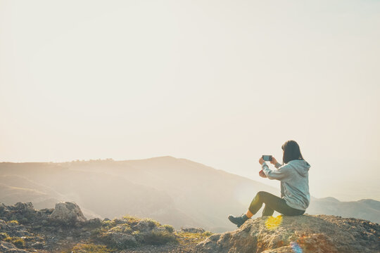 Hipster girl is shooting video of beautiful sunrise on cell telephone, while is standing against bright sky scenery background with copy space. Female tourist is taking photo with mobile phone camera