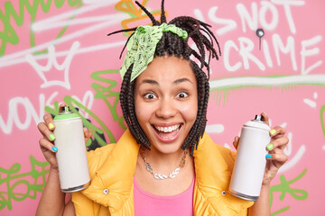 Pretty cheerful Afro American hipster girl with dreadlocks smiles broadly holds two aerosol bottles feels happy and positive uses aerosol sprays for drawing graffiti. Youth art creativity concept - obrazy, fototapety, plakaty