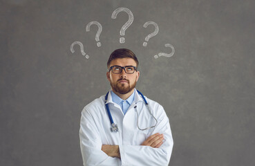 Smart doctor in white lab coat and glasses with question marks above head standing arms folded and...