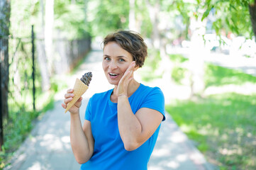 Happy caucasian girl in blue dress eats chocolate ice cream cone outdoors. Emotional excited short-haired woman enjoying a chilling gelato on a hot summer day in the park.