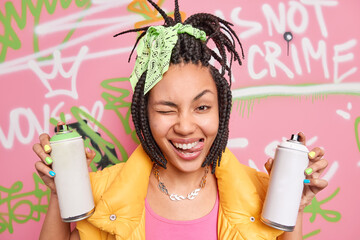 Positive female street artist with trendy hairstyle winks eye sticks out tongue has fun while painting graffiti wall wears clolorful clothes holds aerosol bottles foolishes around. Youth subculture