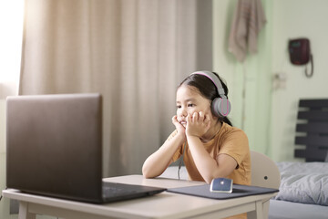 Asian child or kid girl bored e-learning on computer notebook to listening wearing headphone and sit with hand on chin by unhappy study online or people learn from home on laptop by back to school