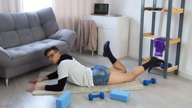 Funny young nerd man in glasses doing fitness exercise lying on floor on carpet and moving legs at home. Training workout sport humor freak comic concept. Newcomer, beginner in sport.