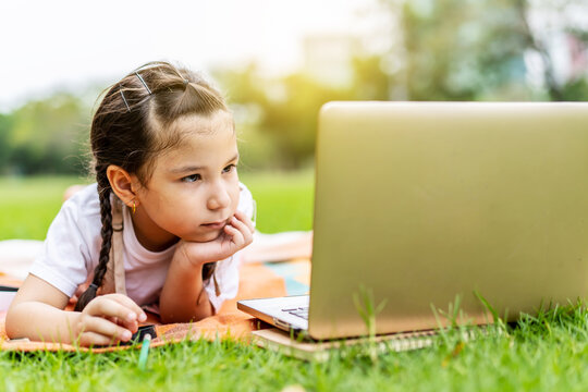 Happy little girl child smiling, talking, learning and having fun looking at laptop computer in summer park. Learning online education concept