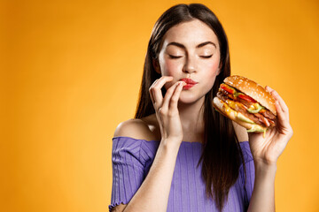Woman eating cheeseburger with satisfaction. Girl enjoys tasty hamburger takeaway, licking fingers...